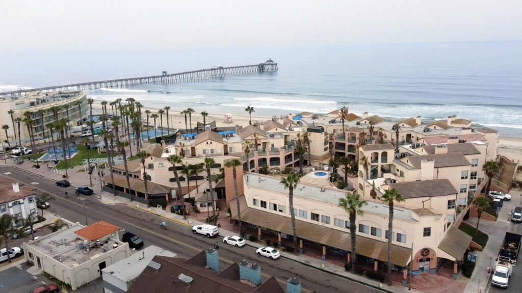 Coastal town with palm trees, residential buildings, and a pier extending into the ocean.